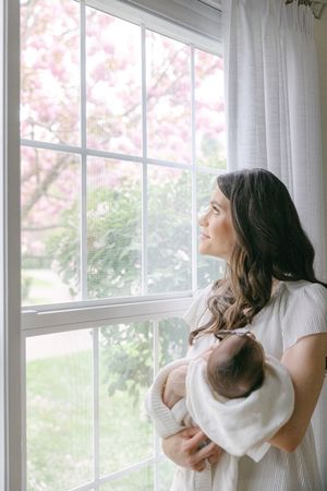 New mother in white dress holds sleeping newborn while gazing out window at blooming pink cherry tree.