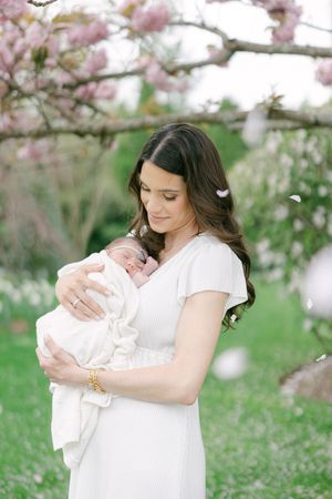 A mother in a white dress tenderly holds her newborn baby under blooming pink cherry blossom trees outdoors.