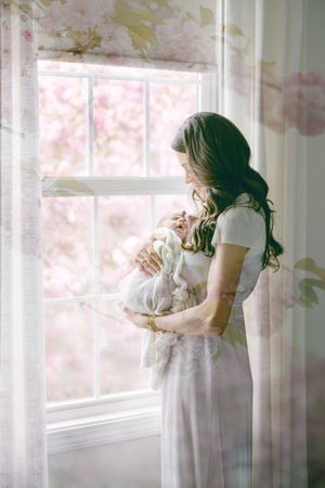 Mother in white dress holds newborn baby by sunlit window with pink floral curtains and blossoming tree outside.