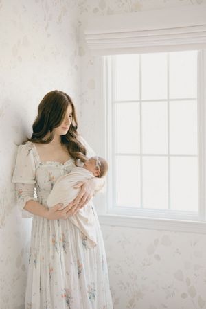 Mother in floral dress tenderly holding swaddled newborn baby by bright window in nursery room.