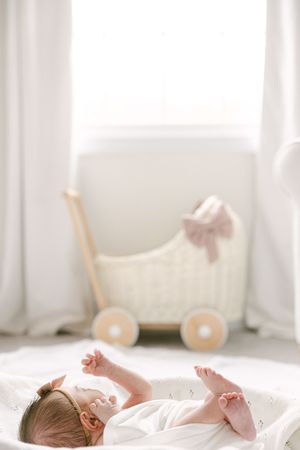 Newborn baby lying on white blanket with tiny toy pram and pink bow in bright nursery room.