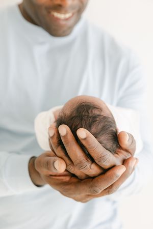 Smiling Black father gently cradling newborn baby's head in his hands, wearing a light blue shirt.