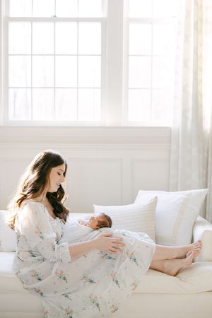 A mother in a floral dress sits on a white sofa, lovingly cradling her newborn baby in soft natural light.