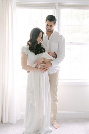 New parents in white outfits tenderly hold their newborn baby by a bright window in a serene home setting.