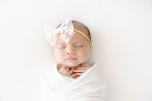 Peaceful newborn baby girl swaddled in white wrap with lace bow headband, sleeping on white background.