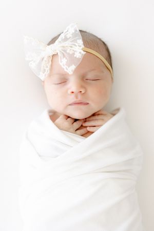 Sleeping newborn baby girl swaddled in white wrap wearing a delicate lace bow headband.