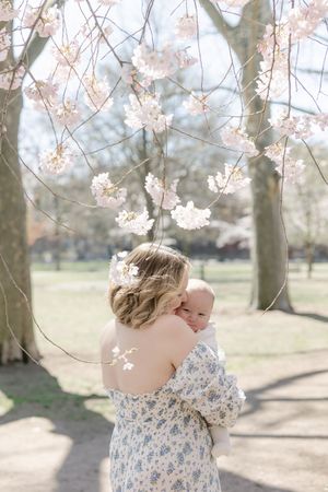 Mother holding baby under cherry blossom tree in spring park, wearing floral dress with pink blooms overhead.