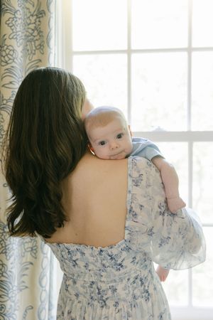 A mother with long dark hair holds her baby against her shoulder near a bright window, wearing a blue floral dress.