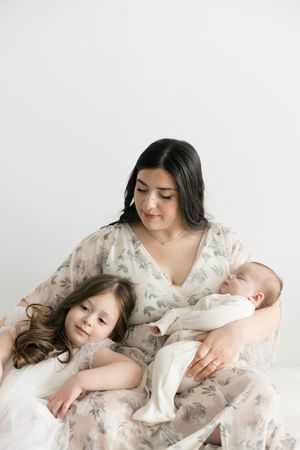 A dark-haired mother in a floral dress tenderly holds her newborn and young daughter in a bright, airy studio.