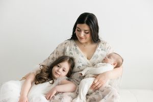 A mother in a floral dress holds her newborn while her young daughter leans against her in a bright studio.