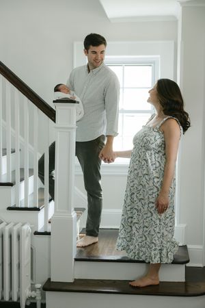 A couple holds hands on a staircase, man carrying newborn baby, woman in floral dress, bright home interior.