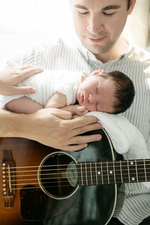 Father holding sleeping newborn baby on acoustic guitar, tender parenting moment with infant in white onesie.