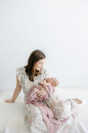 A young girl in a floral dress sits on a bed, lovingly cradling a newborn baby wrapped in a pink blanket.