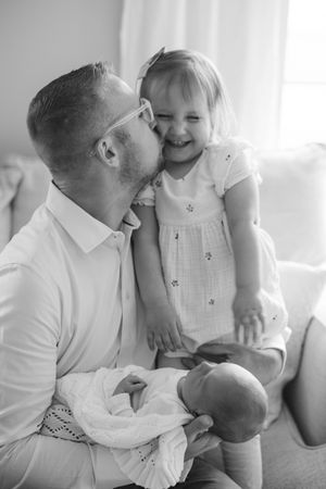 Father kisses laughing toddler daughter while holding newborn baby in black and white photo.