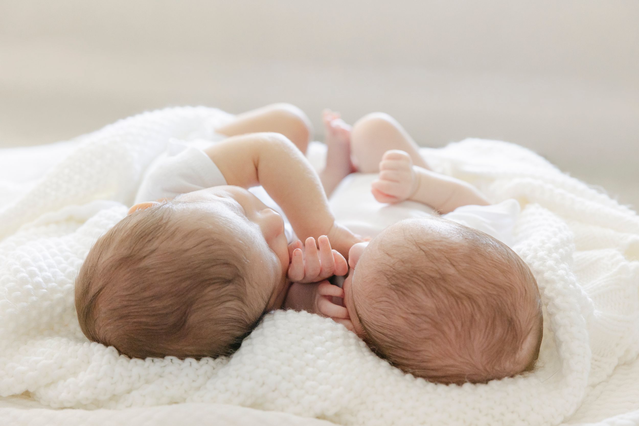 Two newborn twins sleeping peacefully side by side on a soft white blanket, holding hands.