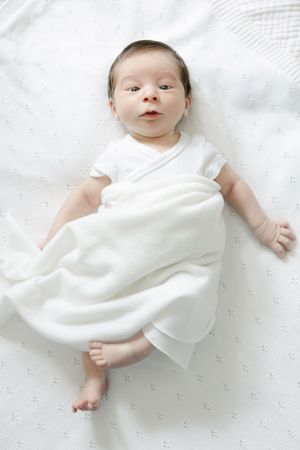 A newborn baby in a white onesie lies on a white blanket, partially swaddled, looking up with bright eyes.
