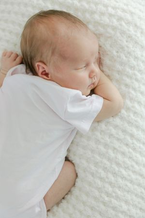 Peaceful newborn baby sleeping on a white knit blanket wearing a white onesie.