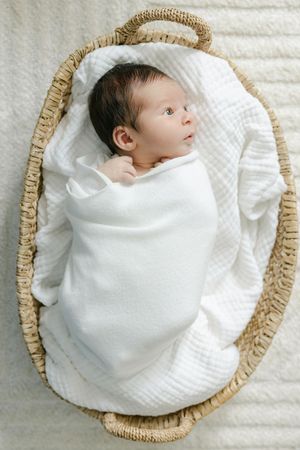Newborn baby swaddled in white blanket lying in a woven Moses basket on a soft cream textured surface.
