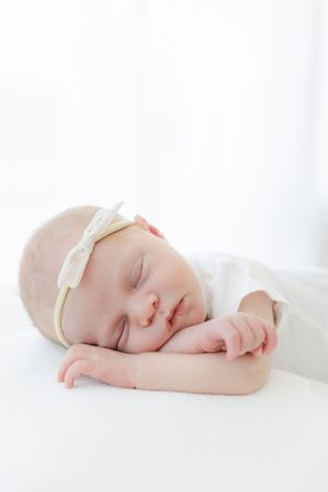 Peaceful newborn baby sleeping with head resting on hands, wearing white bow headband on white background.
