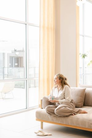 Woman sitting cross-legged on a beige sofa holding a mug, gazing out large floor-to-ceiling windows in a bright modern living room.