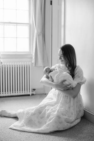 Black and white photo of a mother sitting on the floor cradling her newborn baby by a window.