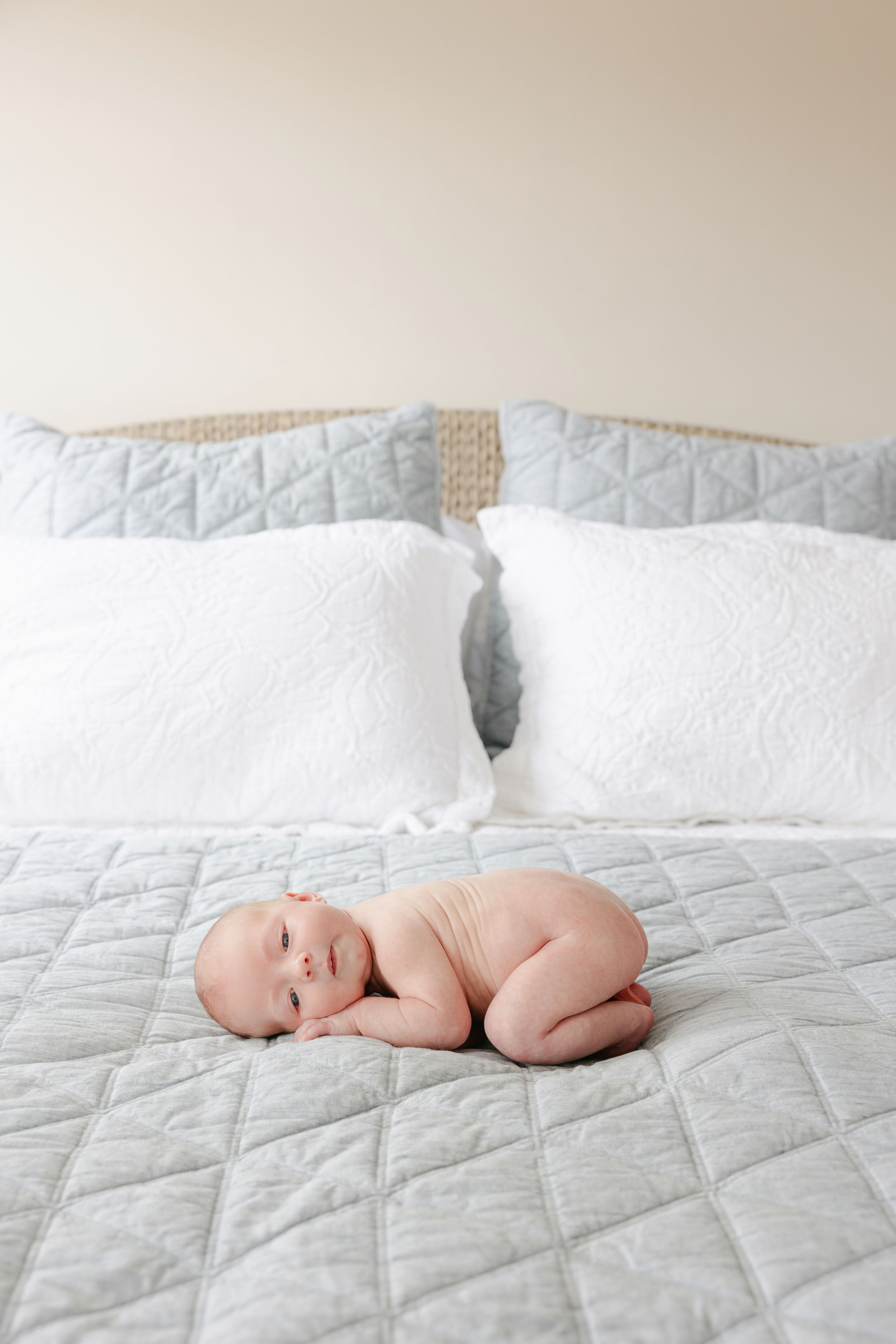 A newborn baby lying on a light blue quilted bedspread with white pillows in the background.
