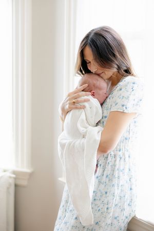 A mother in a floral dress tenderly kisses her newborn baby wrapped in a white knit blanket by a bright window.