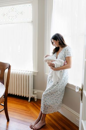 A barefoot mother in a floral dress holds her newborn baby by a bright window with a white radiator.