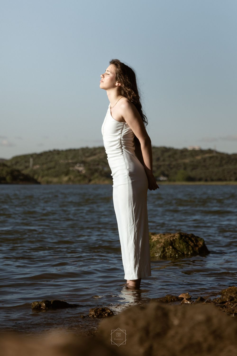Woman in white dress standing in shallow water with scenic lake and green hills in background on a sunny day.
