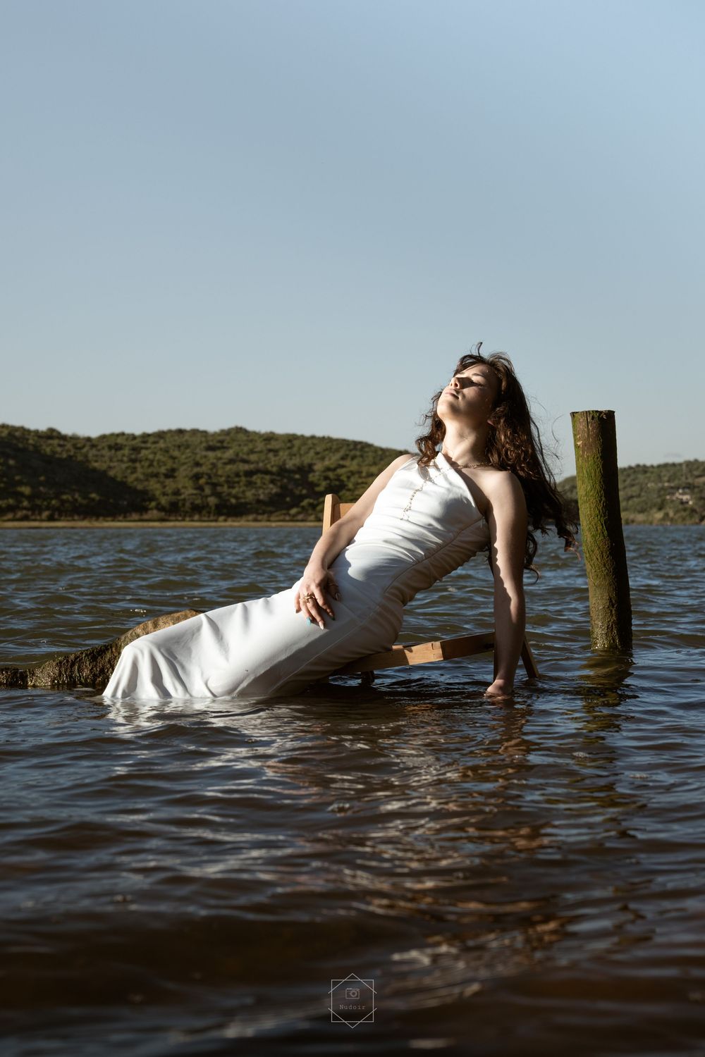 Woman in white dress reclining on submerged chair in lake with lush hills and clear blue sky.