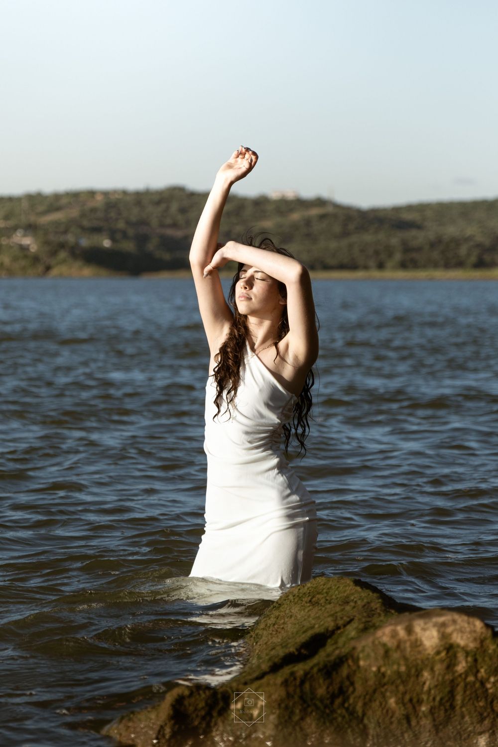 Woman in white dress standing in water with arms raised, rocky shoreline and green hills in background.