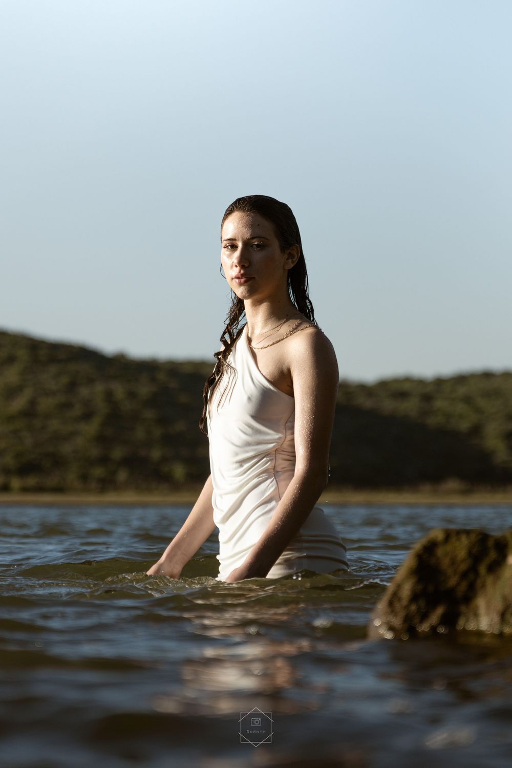 A woman in a wet white dress stands waist-deep in a lake with green hills in the background.