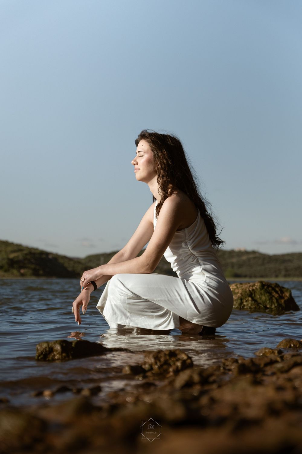 Woman in white outfit crouching by a calm lake, enjoying the serene natural landscape on a sunny day.