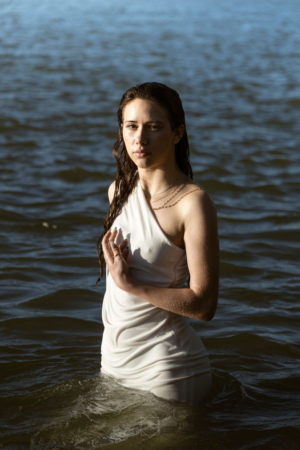 Young woman in wet white dress standing waist-deep in calm water with dark wet hair, looking at camera.