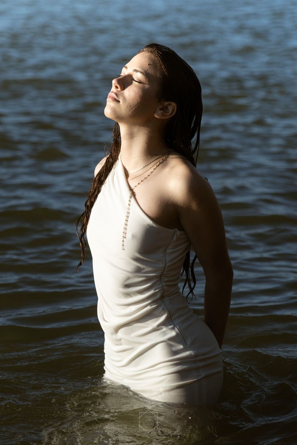 Young woman in wet white dress standing in water with eyes closed, enjoying sunlight on her face.