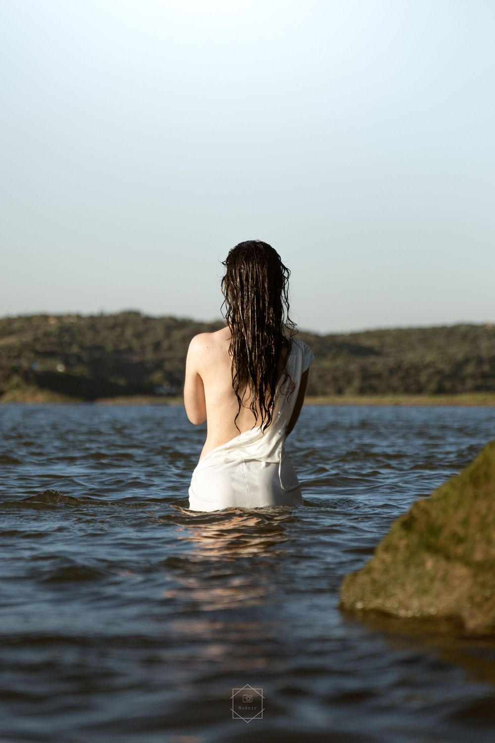 Woman with wet hair standing waist-deep in a calm lake, viewed from behind, wearing a white garment.
