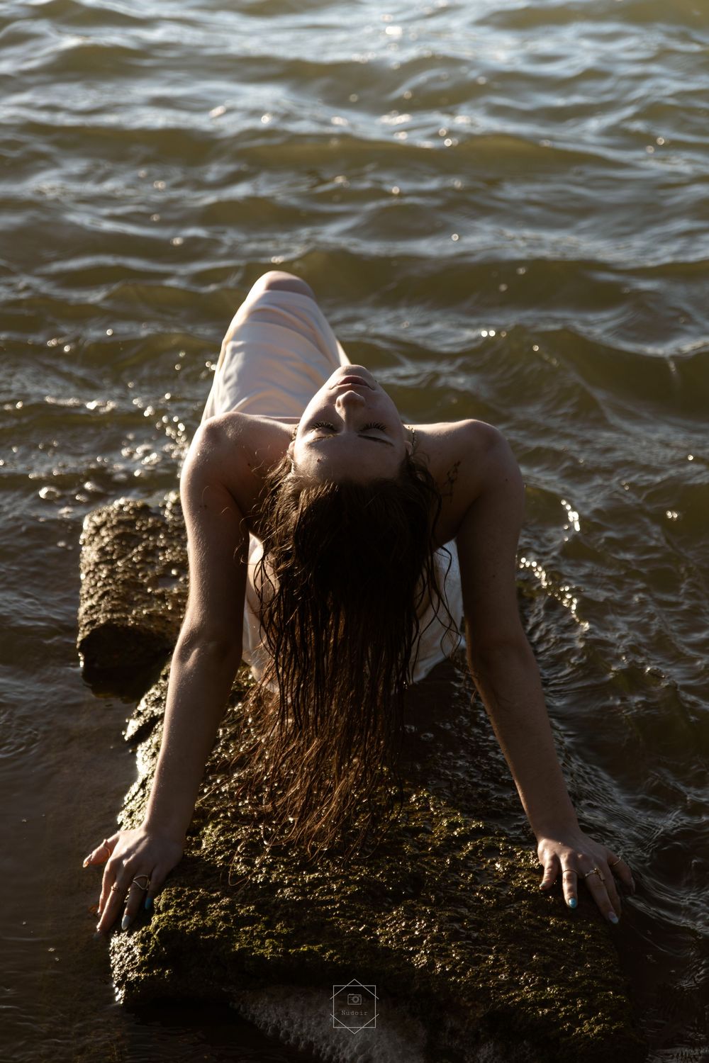 Woman in white dress leaning back on rocks in shallow ocean water, wet hair flowing, golden sunlight reflecting off waves.