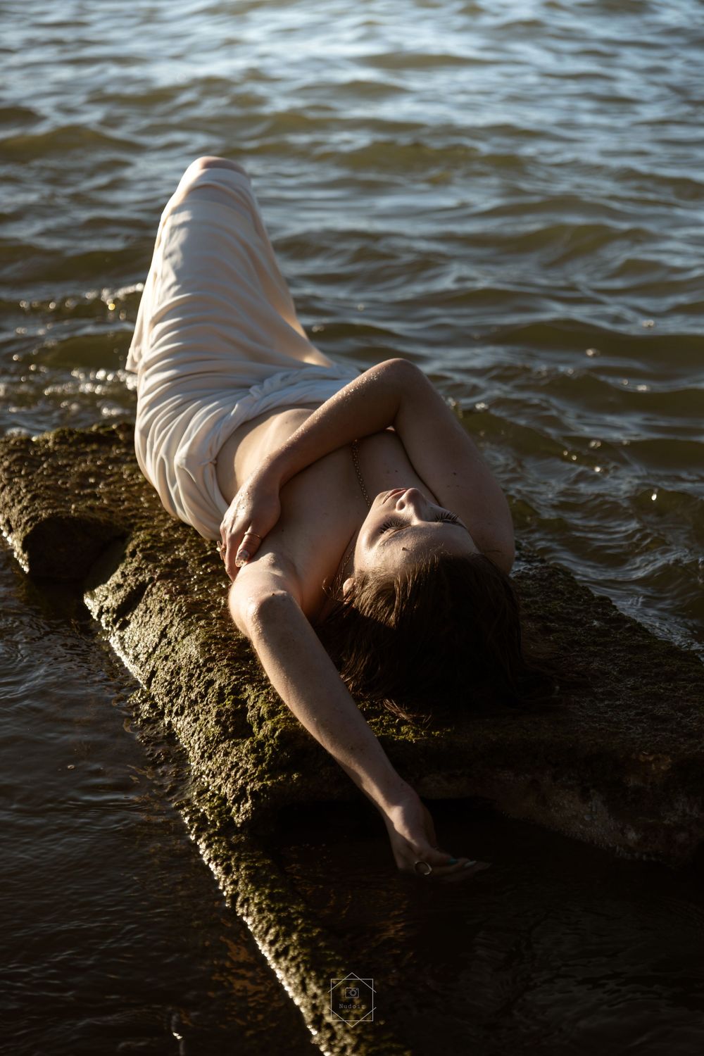 Shirtless man in white pants reclining on mossy rocks at the water's edge, eyes closed in peaceful repose.
