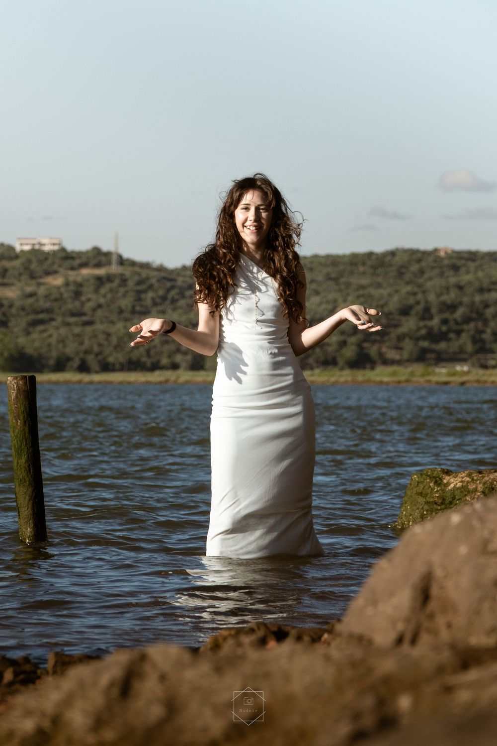 Woman in white dress standing in water with arms outstretched, smiling, with green hills in background.