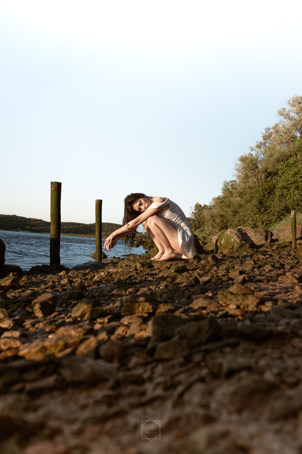 Woman in white dress crouching on rocky shoreline near wooden posts by a calm lake at golden hour.