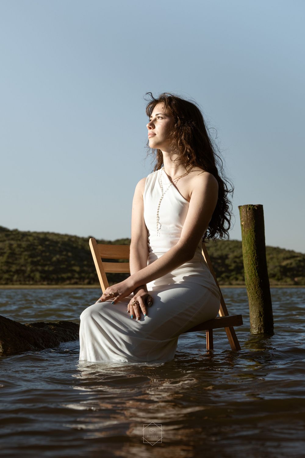 Young woman in white dress sitting on wooden chair in calm lake water with green hills and blue sky behind her.