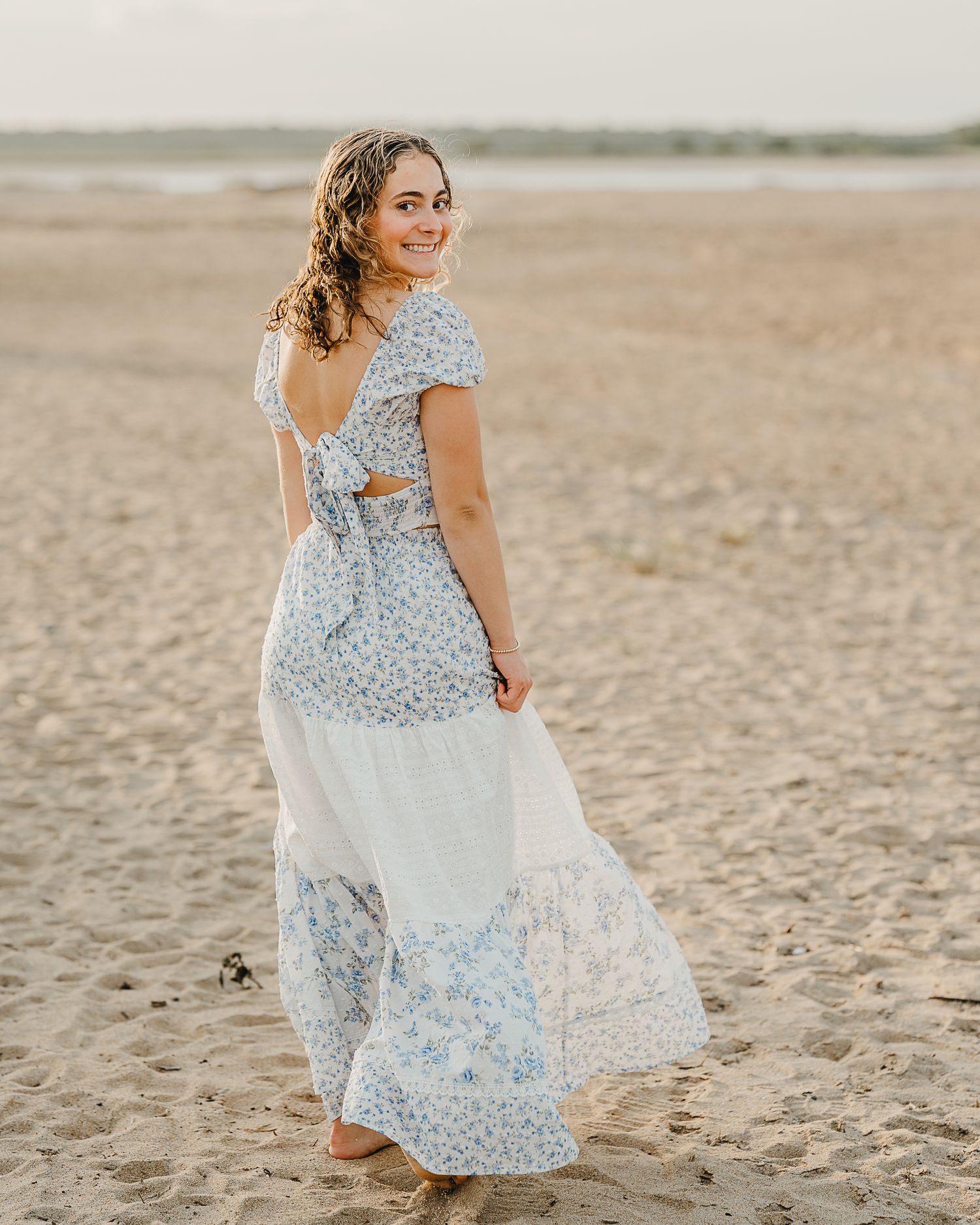 high school senior in long flowy pale pink dress posing on sandy beach at golden hour sunset, smiling with hands in hair.