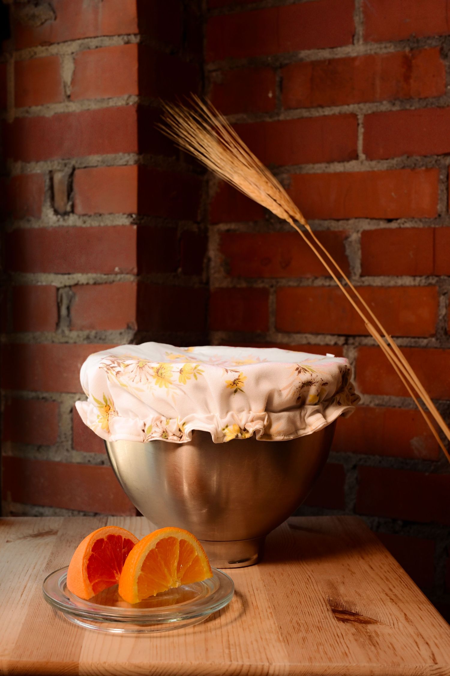 Steel mixing bowl covered with floral cloth near wheat stalks and orange slices on wooden table against brick wall.