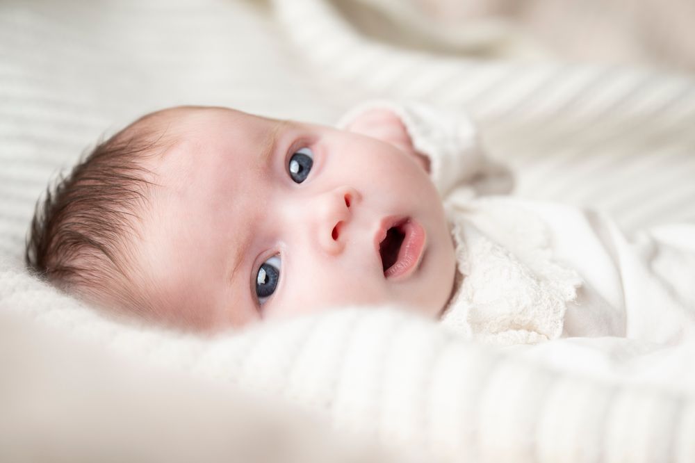 Newborn baby with bright blue eyes lying on soft white bedding, looking up with curiosity – newborn photography in Ashford, Kent.