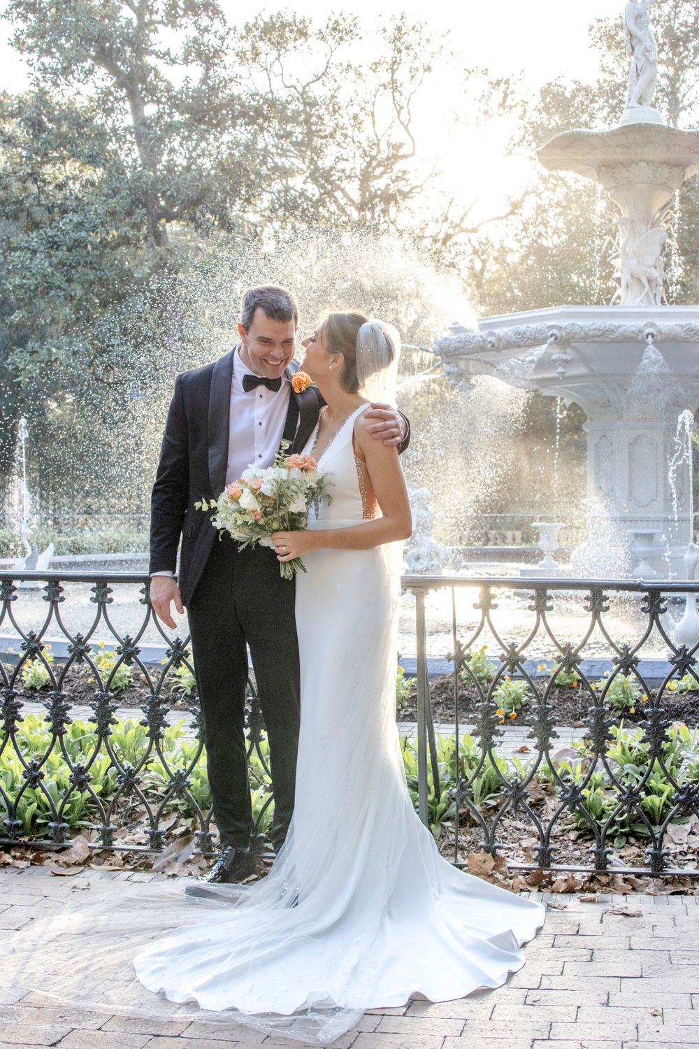 A wedding couple poses by an ornate fountain in a garden setting with beautiful sunlight streaming through the trees.