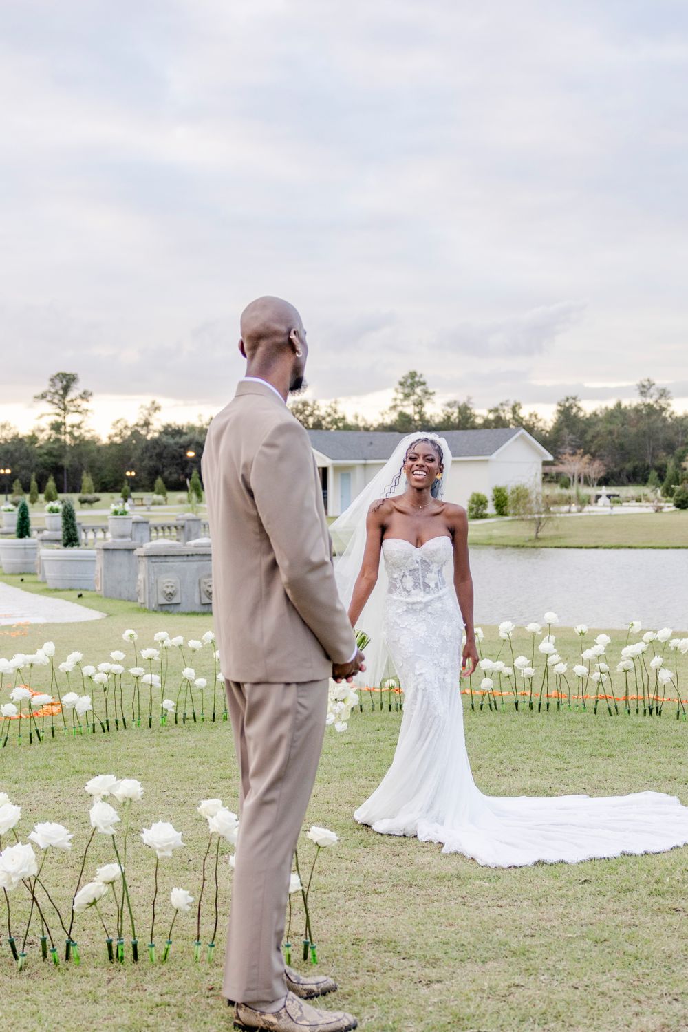 A couple stands in a garden setting with white flower decorations near a lake during golden hour.
