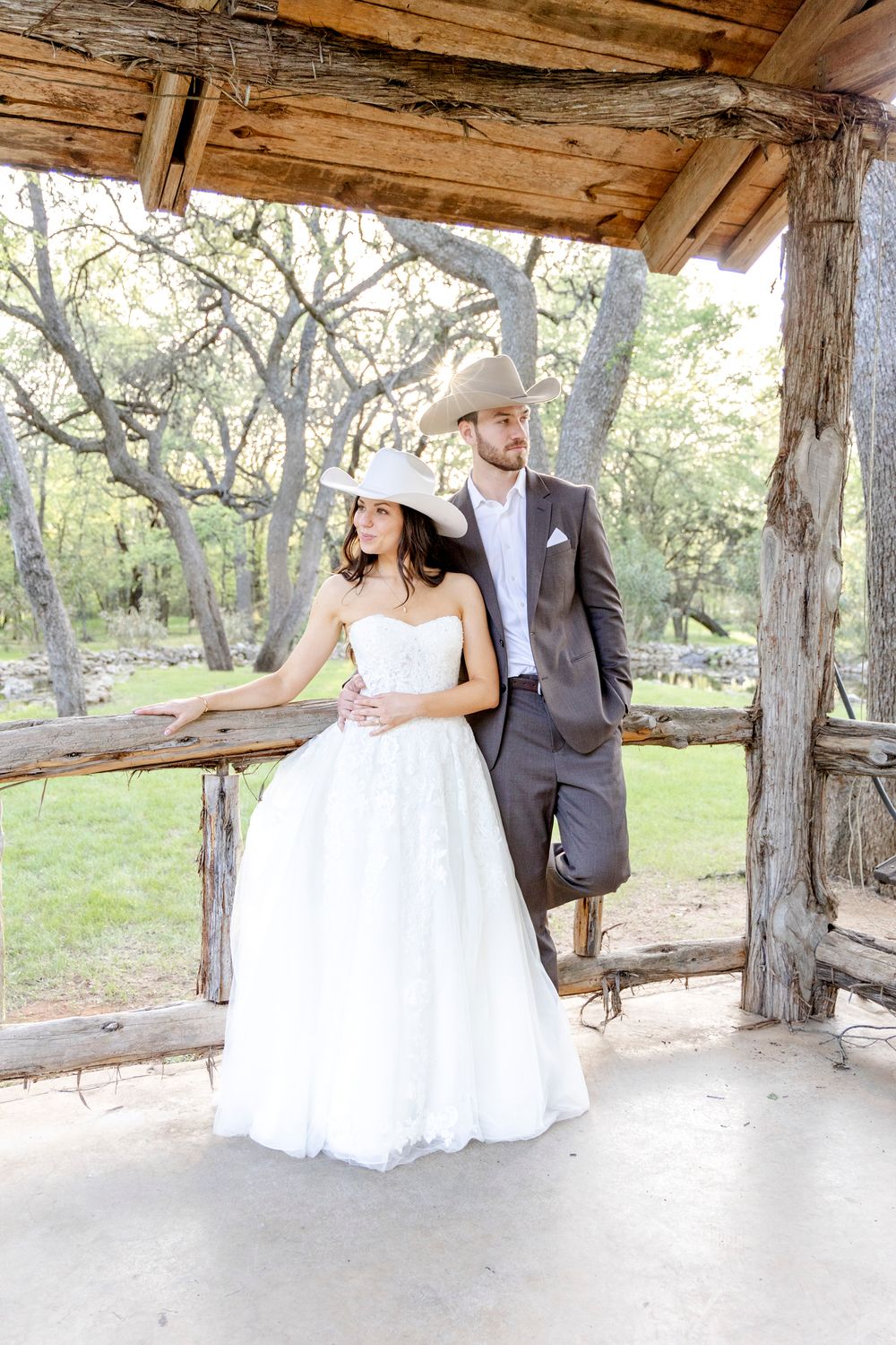 A couple in wedding attire stands under a rustic wooden gazebo surrounded by trees in a natural outdoor setting.