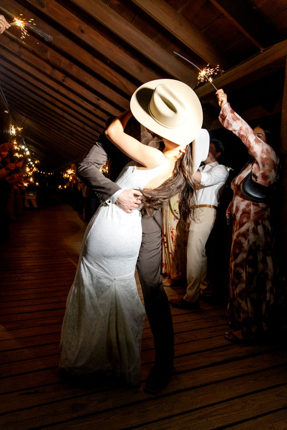 Couple dancing at a rustic wedding reception with string lights overhead and wooden beams visible on the ceiling.