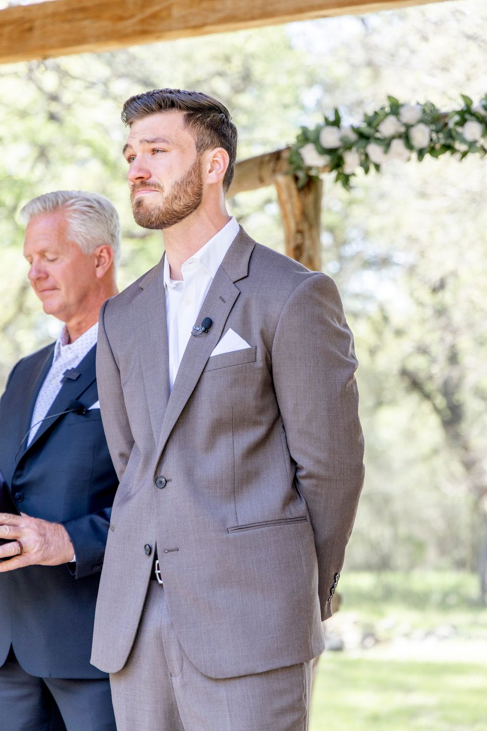 Two people in suits stand under a wooden pergola decorated with floral garlands during an outdoor wedding ceremony.