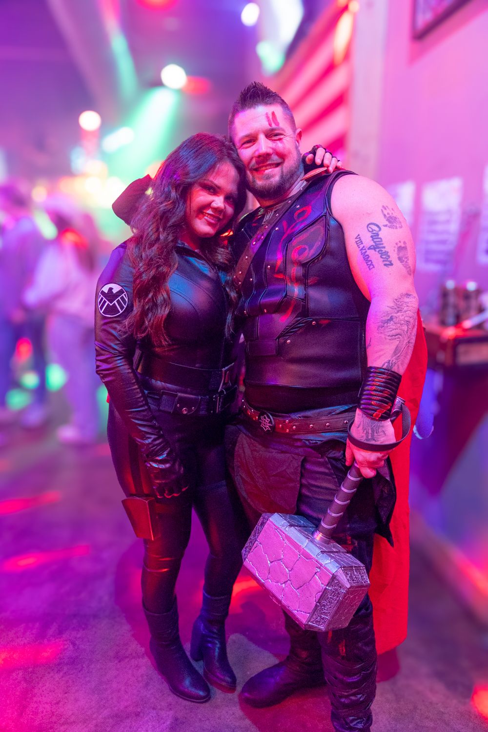 A couple in Marvel costumes, Thor with Mjolnir and an S.H.I.E.L.D. agent, posing at a neon-lit Halloween party.